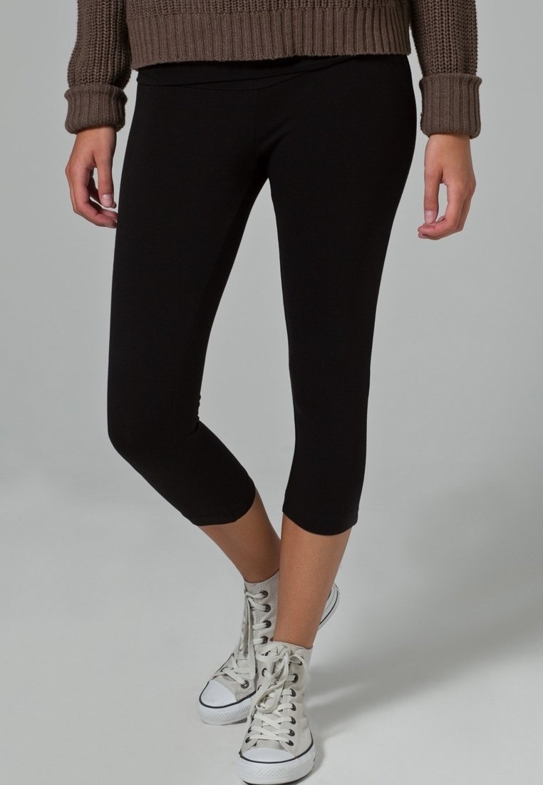Zwarte capri leggings van rekbaar materiaal, met een strakke pasvorm die net onder de knie eindigt, gecombineerd met witte sneakers.