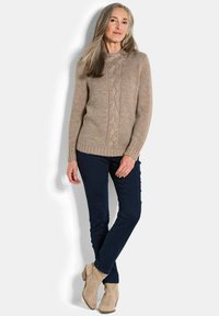 Beige gestrickter Pullover mit einem Zopfmuster, rundem Ausschnitt und geripptem Saum, kombiniert mit dunkelblauen Hosen und beigen Stiefeletten.