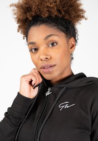 Zwarte rits-hoodie met een zilveren logo, gestructureerde stof, ribbelmanchetten en decoratieve stiksels langs de rits. Persoon is de capuchon aan het verstellen.