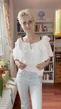 Osoba s krátkými blonďatými vlasy si upravuje bílou volánkovou blůzu, nosí světle modré džíny a stojí před knihovnou v útulném pokoji.
