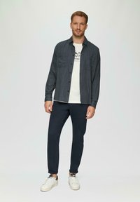 Donkerblauw overhemd met knopen, twee borstzakken, gedragen over een wit T-shirt met opdruk. Gecombineerd met marineblauwe broek en witte sneakers.