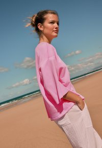 Tricou roz supradimensionat cu mâneci largi, asortat cu pantaloni de culoare deschisă. Textură netedă, croială casual, purtat într-un decor de plajă sub cer albastru.