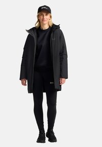 Zwarte geïsoleerde parka met capuchon, gecombineerd met een zwart sweatshirt en leggings. Een zwarte pet heeft een logo aan de voorkant.