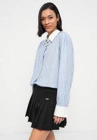 Ανοιχτό μπλε πουκάμισο με κάθετες ρίγες, λευκό γιακά και μανσέτες, συνδυασμένο με μαύρη πλισέ φούστα που φέρει λεπτομέρεια με λογότυπο.