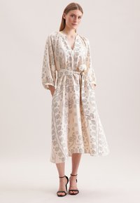 Langes, beiges Kleid mit Paisleymuster, V-Ausschnitt und Dreiviertelärmeln. Inklusive eines Bindegürtels und Seitentaschen. Glattes, leichtes Material.