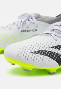 Scarpe da calcio bianche con accenti grigi, logo nero del marchio, tomaia in sintetico testurizzato, suola gialla neon e dettagli a motivo visibili.