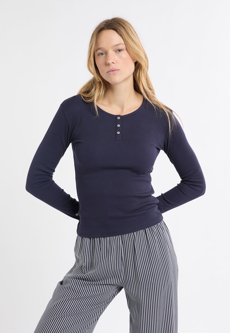 Marineblaues, langärmliges Rippstrick-Oberteil mit rundem Ausschnitt und Drei-Knopf-Leiste. Weiche Textur und figurbetontes Design betonen die Silhouette.