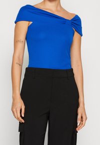 Top azul sin hombros con tela drapeada, diseño ajustado y detalle retorcido en el escote, combinado con pantalones cargo negros de cintura alta.