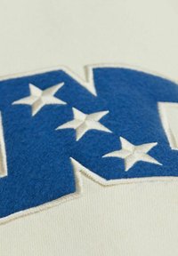 Tessuto bianco con un logo "N" in feltro blu, caratterizzato da stelle bianche. La texture è liscia con bordi ricamati. I dettagli sono chiaramente definiti.