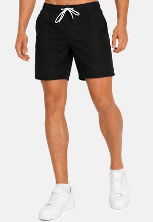 Männliches Modell trägt schwarze Shorts mit Kordelzug und weiße Sneakers, steht vor einem schlichten hellen Hintergrund.