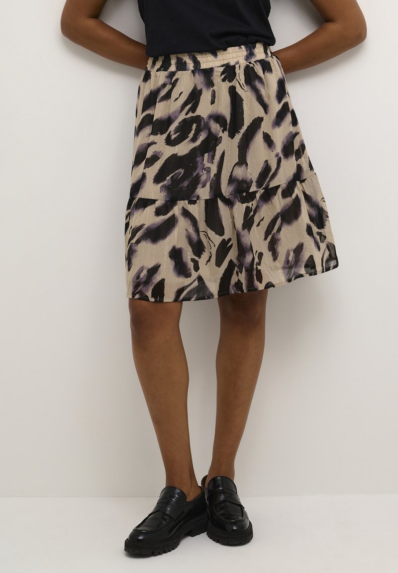 Femme portant une jupe à volants imprimée abstraite beige et noire, un haut noir, et des mocassins noirs épais, debout devant un fond blanc.