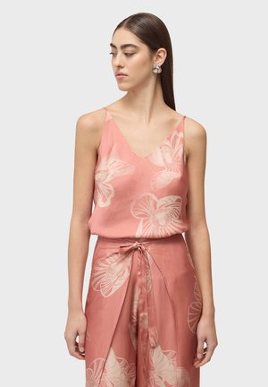 Žena oblečená v ružovom kvetovanom tielku bez rukávov a zodpovedajúcich širokých nohaviciach s detailom viazania vpredu, hľadí doľava.