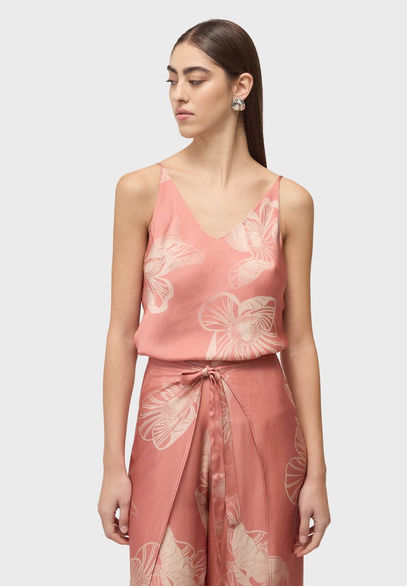 Femme portant un haut sans manches rose à imprimé floral et un pantalon à larges jambes assorti avec un détail noué à l'avant, regardant vers sa gauche.