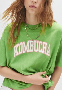 T-shirt verde oversize con la scritta "KOMBUCHA" stampata in bianco e rosa. Tessuto morbido con una finitura leggermente texturizzata. Modello che indossa collane sovrapposte.