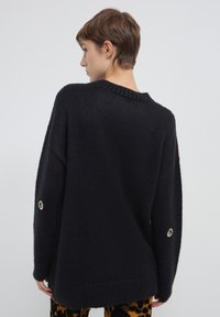 Maglione nero a maglia con vestibilità ampia, collo e polsini a coste, accentuato da occhielli metallici sulle maniche. Morbida texture e design oversize.