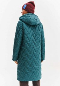 Tyrkysově modrý prošívaný kabát s chevronovým vzorem, kapucí a viditelným logem na bordó pletené čepici, kterou má model na sobě.