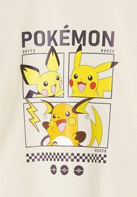 Beige T-Shirt mit einem Grafikdesign, das drei gelbe Pokémon-Charaktere in Comic-Style-Boxen, einen Blitz und schwarze Schachbrettmuster enthält.
