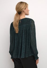 Blusa di un verde scuro con tessuto testurizzato, vestibilità comoda, maniche lunghe con polsini arricciati e dettaglio a chiave sulla schiena. Abbinata a pantaloni neri.