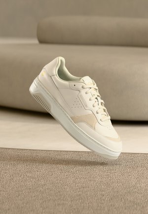 Kremfarget sneaker med snøring og perforert detalj på siden, plassert over et beige teppe og med en sofa i matchende farge i bakgrunnen.