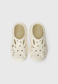 Sabots en caoutchouc beige clair avec une semelle intérieure texturée, des trous d'aération pour la respirabilité, et un design lisse et arrondi. Légers et faciles à nettoyer.