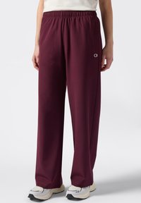 Persoon in ruime bordeauxrode sweatpants en witte sneakers, staand tegen een effen witte achtergrond.