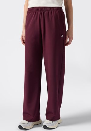 Persoon in ruime bordeauxrode sweatpants en witte sneakers, staand tegen een effen witte achtergrond.