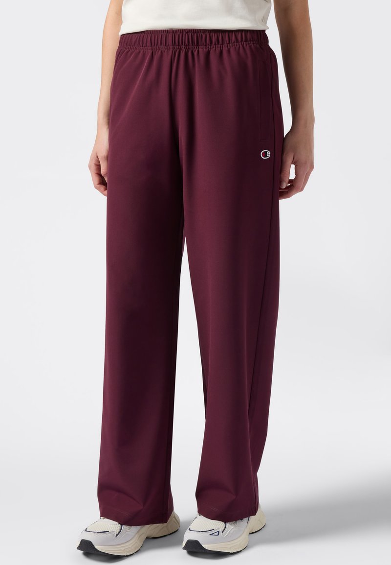 Persoon in ruime bordeauxrode sweatpants en witte sneakers, staand tegen een effen witte achtergrond.