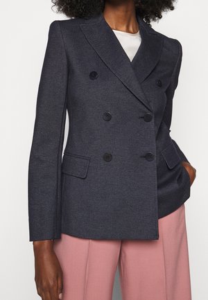 Kvinde iført en tætsiddende mørkegrå dobbeltradet blazer med sorte knapper og pink bukser med brede ben, hånd i lommen.