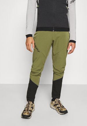 Homme portant un pantalon de sport olive vert et noir avec le logo "DYNAFIT" et des chaussures de course sur sentier beiges, debout devant un fond clair uni.