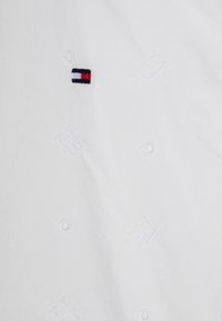 Biała tkanina z subtelnym tłoczonym wzorem z okrągłymi i geomerycznymi motywami. Na tkaninie znajduje się małe haftowane logo w granatowym, czerwonym i białym kolorze.