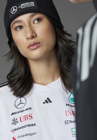 Camiseta deportiva blanca con acentos negros, que presenta los logotipos de Mercedes, AMG y marcas patrocinadoras. Gorro de punto negro con detalle del logotipo.