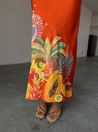 En livlig oransje kjole har tropiske frukter og plante-mønstre ved sømmen, og er kombinert med sorte stroppesandaler på bare føtter.