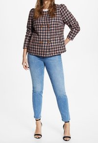 Chaqueta de corte cruzado en patrón de espiga en rosa oscuro y negro, con dos botones grandes, combinada con jeans ajustados de color azul claro y tacones negros.