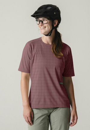 Frau mit schwarzem Fahrradhelm, klarer Brille, kurzärmeligem mauve-karierter Hemd und hellgrauer, perforierter Hose, die lächelnd nach rechts schaut.