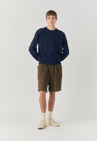 Felpa blu navy con collo a giro, abbinata a pantaloni cargo color khaki con due tasche laterali e cintura elastica, indossati con sneakers bianche.