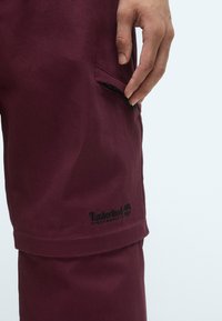 Bordeauxfarbene Shorts aus strukturiertem Stoff, mit einer Seitentasche und einem schwarz gestickten Timberland-Logo nahe dem Saum.
