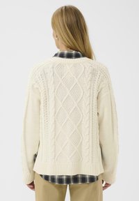Un cardigan crème en tricot à mailles torsadées présentant un motif en losanges au dos, des poignets côtelés et une chemise à carreaux contrastante en dessous.