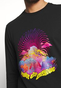 Schwarzes Langarmshirt mit buntem psychedelischem Bergdesign und gelbem Text "Never Stop Exploring" in der Mitte unten.