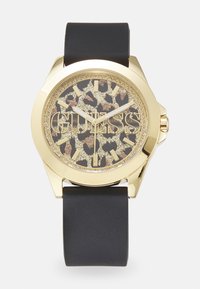 Montre en or avec un bracelet en silicone noir. Le cadran rond présente un motif léopard et le logo doré "GUESS", avec des index et aiguilles dorés.