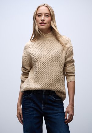 Pull en tricot beige avec un col montant, présentant des motifs de torsades texturés et des manches ajustées, associé à un jean bleu foncé.