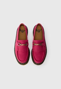 Pink leather loafers with a black sole, featuring a fringe detail and a metallic buckle accent. Textured upper and stitched edges.