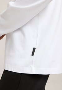 Biała koszula z długim rękawem o zaokrąglonym dolnym wykończeniu, z czarnym detalem metki po boku. Wykonana z miękkiej bawełnianej tkaniny o gładkiej fakturze.