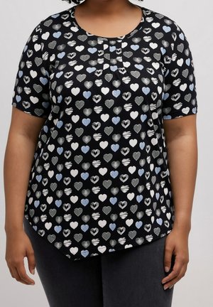 Femme portant un tee-shirt noir à manches courtes orné de cœurs blancs, bleus et gris, associé à un pantalon sombre, se tenant devant un fond uni.