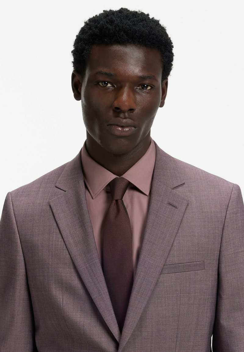 Lichtpaarse colbert, bijpassend overhemd en een donkere stropdas. Het pak heeft een slank ontwerp, een gestructureerde stof en een detail aan de voorkant van de zak.