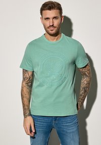 Zelené bavlněné tričko s kulatým výstřihem a embosovanou kruhovou grafikou tenisu. Nosené s modrými džínami, s tetováním na ruce.