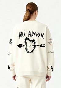 Felpa color crema con grafiche nere e il testo "Mi Amor", teschi, cuori, frecce e un motivo a saette. Vestibilità casual e oversize.