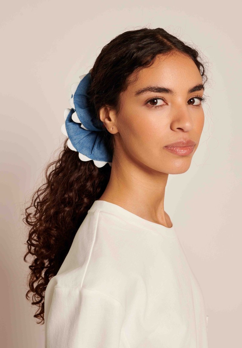 Scrunchie de denim com destaques brancos em forma de ondas, tecido texturizado e formato circular, usado em um rabo de cavalo solto contra um fundo neutro.