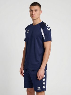 Młody mężczyzna stoi, ubrany w granatową odzież sportową z białymi wzorami w kształcie litery V na ramionach i szortach, neutralny wyraz twarzy, jednolite tło.