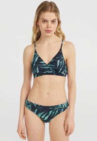 Giovane donna che indossa un bikini scuro con motivi di foglie verdi e teal, in piedi su uno sfondo bianco semplice.