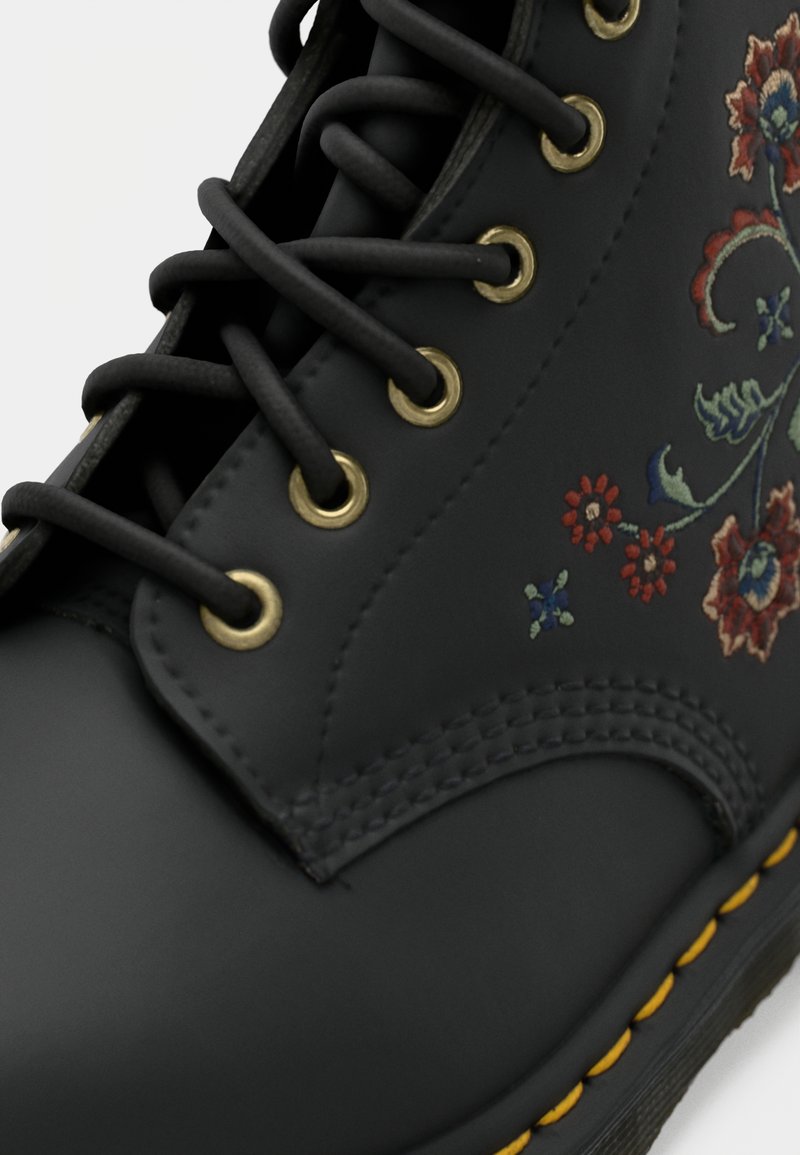 Botte en cuir noir avec lacets noirs, œillets en laiton, couture de semelle jaune, et broderie florale multicolore sur le côté.